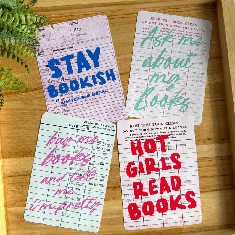 May include: Four rectangular cards with text on a wooden tray. The cards have a library card design with phrases like "Stay Bookish," "Ask me about my books," "Buy me books and tell me I'm pretty," and "Hot Girls Read Books."