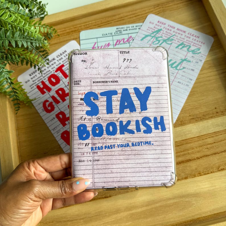 May include: A clear phone case featuring a vintage library card design with the words "STAY BOOKISH" in blue. The case is surrounded by library-themed cards with phrases like "Ask me out" and "Hot Girl Reads."