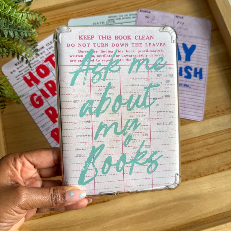 May include: Clear phone case with a vintage library card design. The case features the text "Ask me about my books" in teal script. Other library cards are visible in the background.