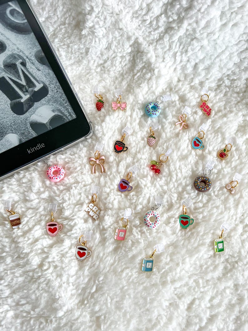 May include: A collection of colorful enamel charms with various designs, including donuts, coffee cups, books, strawberries, hearts, and bows. The charms are attached to clear plastic clips, which can be used to mark pages in a book or journal.