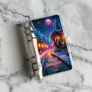 May include: A clear plastic A6 binder with a six-ring metal mechanism. The cover features a vibrant illustration of a path leading to a house, with candy cane borders, glowing street lamps, a full moon, and the text Candy Cane Lane.