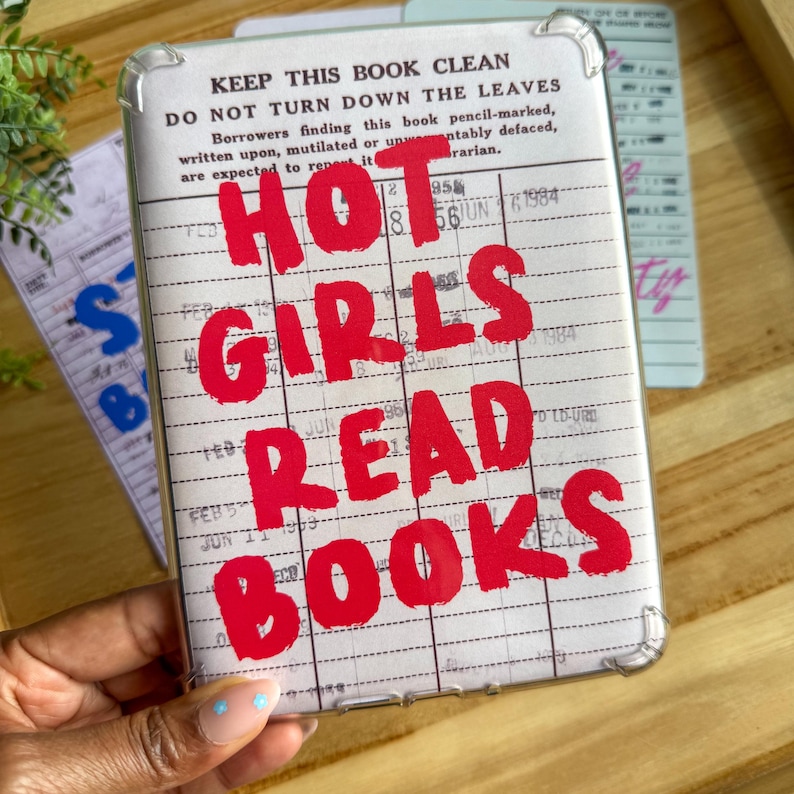 May include: Clear phone case with a vintage library card design. Bold red text reads "HOT GIRLS READ BOOKS" over a background of lines and text. The case has a clear border and is held by a hand.