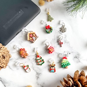 May include: A collection of Christmas-themed charms, including a gingerbread house, Christmas tree, wreath, snowflake, stocking, candy cane, snowman, and penguin. The charms are attached to clear clips, suitable for use with an e-reader. The background features pine cones and gold ornaments.