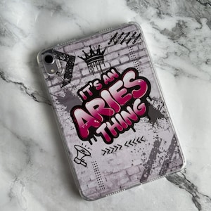 May include: Clear phone case with a graffiti-style design. The case features a brick wall background with a crown, paint splatters, and the text "IT'S AN ARIES THING" in pink and white. Additional graphic elements include a megaphone and arrows.