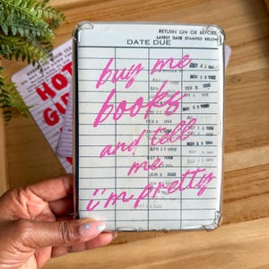 May include: A clear phone case with a white insert resembling a library card. The pink text reads "buy me books and tell me I'm pretty." The case is held by a hand with a light blue nail design.