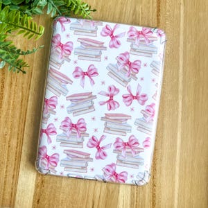 Può includere: Una custodia trasparente per tablet con sfondo bianco e un motivo di fiocchi rosa e pile di libri.