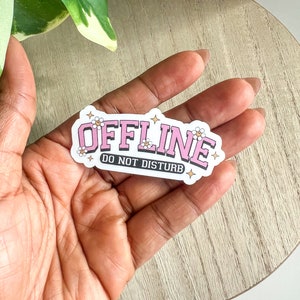 May include: Pink and white sticker with the text "OFFLINE DO NOT DISTURB" in a retro font. The sticker has small white flowers and gold stars around the text.