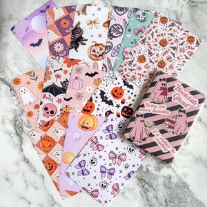 Può includere: Una collezione di segnalibri a tema Halloween e una copertina di libro. I segnalibri presentano vari motivi tra cui zucche, pipistrelli, fantasmi, teschi e fiocchi. La copertina del libro ha strisce rosa e nere con illustrazioni di fantasmi.