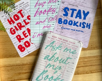 Op de afbeelding: Een verzameling items met bibliotheekthema, waaronder kaarten en een telefoonhoesje. De kaarten bevatten zinnen als "Hot Girls Read Books", "Buy me books" en "Stay Bookish". Op het telefoonhoesje staat "Ask me about my books".