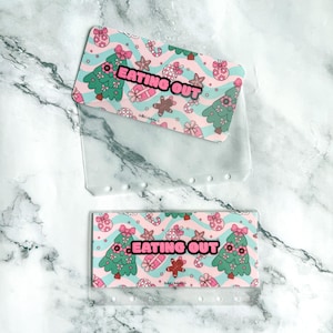 May include: Two rectangular clear plastic cash envelopes featuring a repeating Christmas pattern of green trees, pink ornaments, and gingerbread men on a wavy light blue background, with the text EATING OUT printed in bold pink letters.