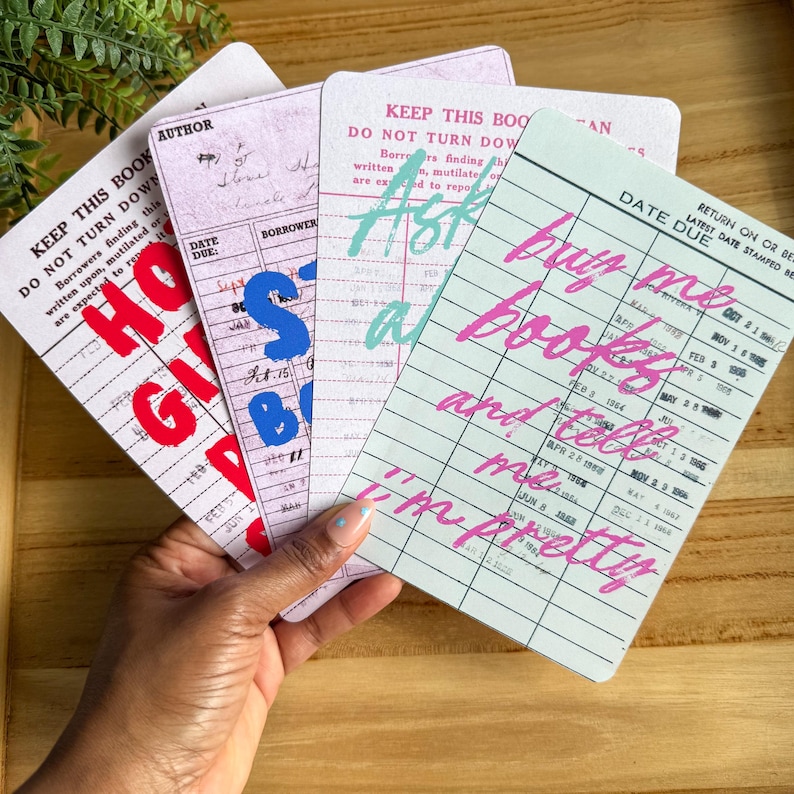 May include: A hand holds a stack of vintage-style library cards. The cards feature text in red, blue, and teal, with phrases like "HO GIB," "Ask a," and "buy me books and tell me I'm pretty." The cards have a retro aesthetic.