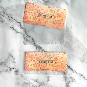 May include: Two clear plastic cash envelopes for a binder, each featuring a top section with an orange and yellow autumn leaf pattern and the text Eating Out printed in a rectangular frame. The envelopes have six punched holes along the bottom.