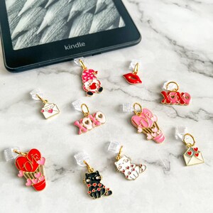May include: A collection of 10 Valentine's Day themed bookmark charms. The charms feature a variety of designs, including a gnome, a heart, a hot air balloon, a cat, a letter, and a lips. The charms are made of metal and have a gold finish.