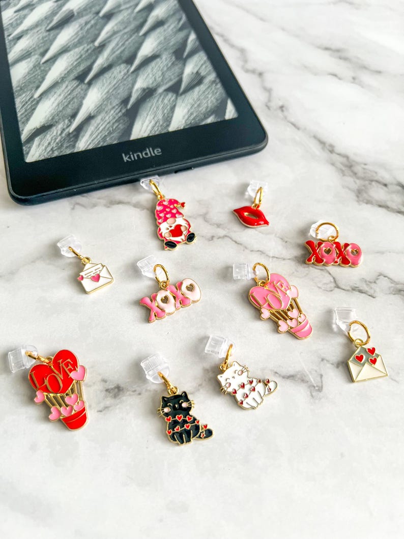May include: A collection of 10 Valentine's Day themed bookmark charms. The charms feature a variety of designs, including a gnome, a heart, a hot air balloon, a cat, a letter, and a lips. The charms are made of metal and have a gold finish.