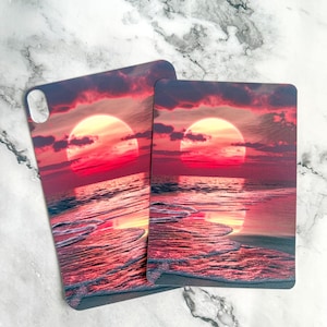 May include: Two rectangular protective cases featuring a vibrant sunset scene over the ocean. The design displays a large, glowing sun in shades of red and pink, with dark clouds and light reflecting across the water and onto the sandy shore.
