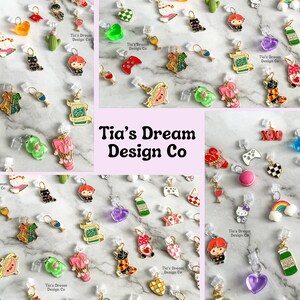 May include: Assortment of colorful enamel charms with various designs, including hearts, animals, food, and gaming-themed charms. The charms are attached to clear plastic clasps. The charms are arranged on a white surface with a green and white patterned background. The text "Tia's Dream Design Co" is visible in the center of the image.
