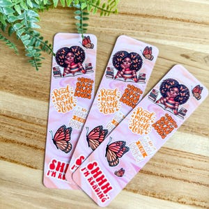 May include: Three pink bookmarks with illustrations of a girl reading, butterflies, and text. The text includes "Read more, scroll less," "My weekend is all booked," and "Shhh, I'm reading."