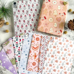May include: A selection of phone and tablet cases with festive designs. The cases display patterns of dogs in Santa hats, gingerbread cookies, and holiday-themed illustrations. The colour palette includes pink, white, and red.