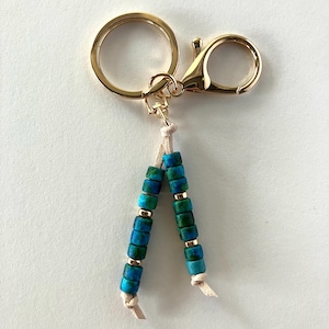 May include: A gold key chain with a blue and green beaded tassel. The beads are small and round, and they are strung on a thin cord. The key chain is attached to a gold ring and a gold clasp.