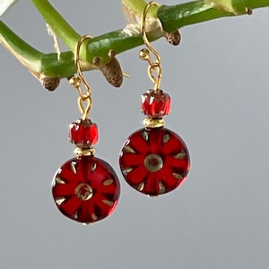 May include: A pair of red and gold earrings. Each earring has a red, circular pendant with a floral design, accented with gold details. The earrings are suspended from gold-coloured ear wires.
