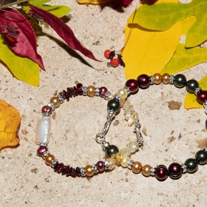 May include: Two bracelets with a mix of gold, red, and green beads. One bracelet has a large white pearl. The bracelets are on a background of fall leaves.