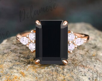 Könnte beinhalten: Ein Verlobungsring aus Roségold mit einem großen schwarzen Smaragdschliff-Edelstein und kleineren klaren runden Edelsteinen auf der Ringschiene.