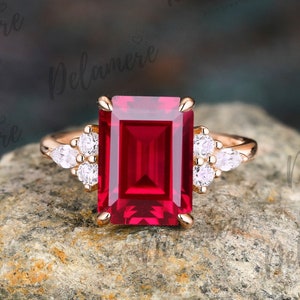 May include: A rose gold ring featuring a large emerald cut ruby gemstone and smaller round diamonds on the band.