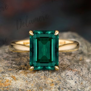 Könnte beinhalten: Ein goldener Ring mit einem großen smaragdgrünen Edelstein im Smaragdschliff. Der Ring liegt auf einem rauen, grauen Stein.
