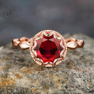 May include: A rose gold ring featuring a large, round, faceted, deep red gemstone. The gemstone is set in a decorative bezel. The band has a leaf-like design with small accent stones. The ring is displayed on a textured grey rock.