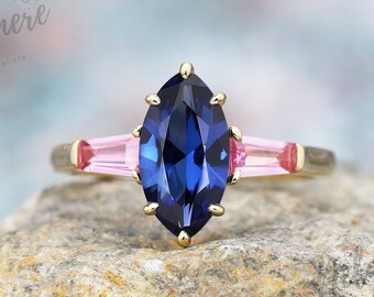 May include: A gold ring featuring a large, marquise-cut, deep blue gemstone as the centerpiece, flanked by two pink baguette-cut stones. The ring is set on a natural stone surface, with the Delamere Fine Jewellery logo visible in the background.