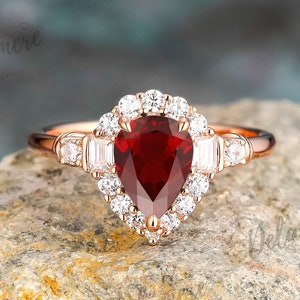 May include: A pear-shaped ruby engagement ring set in rose gold with a halo of smaller diamonds. The ring is on a rough, brown rock.