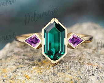 May include: A gold ring with a large, emerald green gemstone in a hexagonal setting. Two smaller, purple gemstones are set on either side of the main stone.