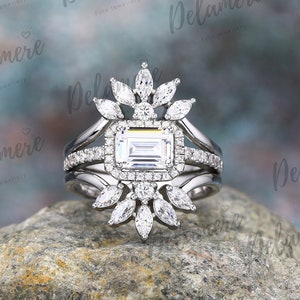 May include: A three-piece engagement ring set with a central emerald-cut diamond and a halo of smaller stones. Marquise-cut diamonds create a floral pattern. The ring, set on a silver band, is displayed on a rock. The brand name "Delamere" is visible.