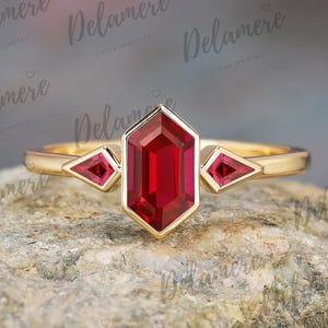 May include: A gold ring featuring a large, rectangular, red gemstone in the center, flanked by two smaller, triangular red gemstones. The ring has a delicate band and is set against a textured, neutral background.