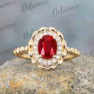 Puede incluir: Un anillo de oro con una gran piedra de rubí ovalada rodeada por un halo de diamantes más pequeños. El anillo tiene un diseño delicado y ornamentado.