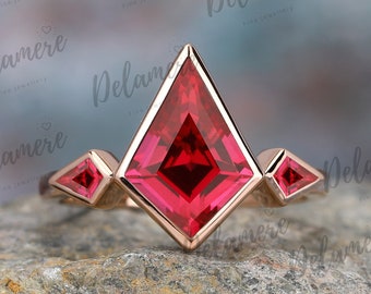 May include: A rose gold ring with a large, square-shaped ruby gemstone in the center. The ruby is surrounded by three smaller, triangular ruby gemstones. The ring is set on a gray, textured surface.