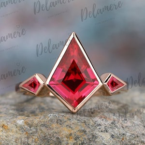 May include: A rose gold ring featuring a large, square-shaped ruby gemstone in the centre. The ruby is surrounded by three smaller, triangular ruby gemstones. The ring is set on a grey, textured surface.