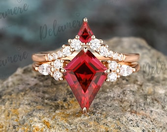 May include: A rose gold engagement ring set with a large, red, square-shaped gemstone surrounded by smaller, clear gemstones. The ring is on a gray, rocky surface.