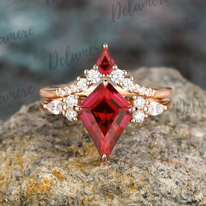 May include: A rose gold engagement ring set with a large, red, square-shaped gemstone surrounded by smaller, clear gemstones. The ring is on a grey, rocky surface.