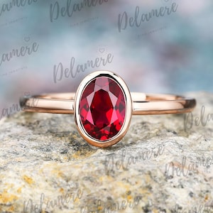 Könnte beinhalten: Roségoldener Ring mit einem oval geschliffenen, leuchtend roten Edelstein. Der Edelstein ist in einer Zargenfassung gefasst, was ein glattes, poliertes Aussehen erzeugt. Der Ring wird auf einer strukturierten, grauen Steinoberfläche gezeigt. Feiner Schmuck.