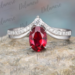 Può includere: Anello in argento con un grande rubino ovale. L'anello presenta una fascia curva con piccole pietre preziose bianche. Una pietra preziosa bianca più piccola si trova sopra il rubino. L'anello è esposto su una superficie grigia strutturata.