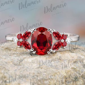 May include: A silver ring with a large, oval-cut, deep red gemstone at its centre. It's flanked by smaller, petal-shaped red gems and accent diamonds. The ring is set on a textured, light-coloured surface. The brand name "Delamere Fine Jewellery" is visible.