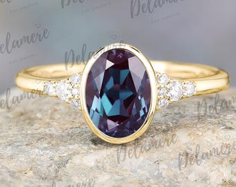May include: A gold ring featuring an oval-cut alexandrite gemstone with a teal and purple color. The ring is accented with small, round diamonds on each side of the main stone. The ring is set on a stone surface.