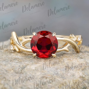 May include: A gold ring with a large, round, red gemstone as the centerpiece. The band features a nature-inspired design with delicate branches and small accent stones. The ring is set on a textured, gray surface.