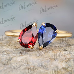 Könnte beinhalten: Ein goldener Ring mit zwei tropfenförmigen Edelsteinen, einem roten und einem blauen. Die Edelsteine sind nebeneinander angeordnet. Der Ring wird auf einer strukturierten, hellen Oberfläche präsentiert.