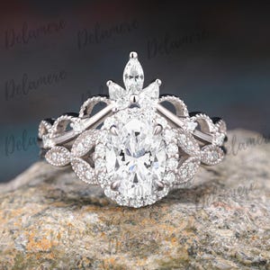 Puede incluir: Un anillo de compromiso de diamantes con una piedra central de talla ovalada, rodeada por un halo de diamantes más pequeños. La banda presenta detalles intrincados y diamantes adicionales. El anillo está sobre una superficie de piedra.