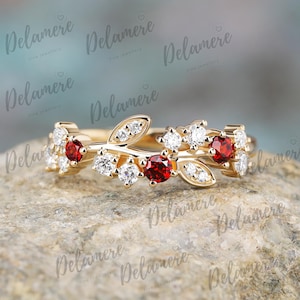 May include: A gold ring with a leaf and branch design. The ring features red and white gemstones. The gemstones are round and set in the gold band. The ring is on a stone surface.