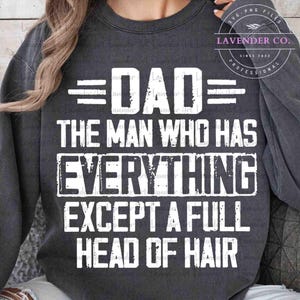 May include: Dark gray sweatshirt with white text that reads "DAD THE MAN WHO HAS EVERYTHING EXCEPT A FULL HEAD OF HAIR." The text is in a bold, sans-serif font. The sweatshirt has long sleeves and a crew neck.