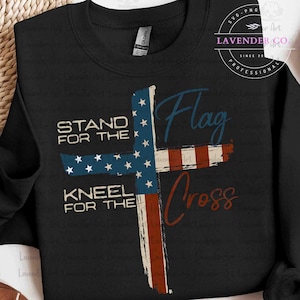 Może przedstawiać: Czarna bluza z patriotycznym krzyżem. Krzyż jest zaprojektowany jak flaga amerykańska, z niebieskimi gwiazdami i czerwono-białymi paskami. Tekst na koszulce brzmi "Stand for the" i "Kneel for the" z "Flag Cross" kursywą.