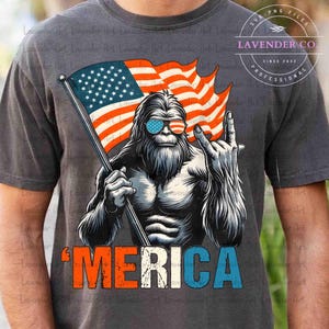 Puede incluir: Camiseta gris oscuro con un dise&ntilde;o de Bigfoot con gafas de sol con la bandera estadounidense, sosteniendo una bandera estadounidense y haciendo el gesto de rock. La palabra "'MERICA" est&aacute; impresa en rojo, blanco y azul. La camiseta tiene el texto "LAVENDER CO" y "SINCE 2022".